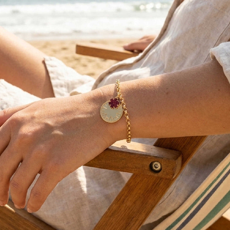 A gold chain bracelet with a round gold medical ID medallion and a small red star of life charm, worn on a woman's wrist in a beach chair at the beach.