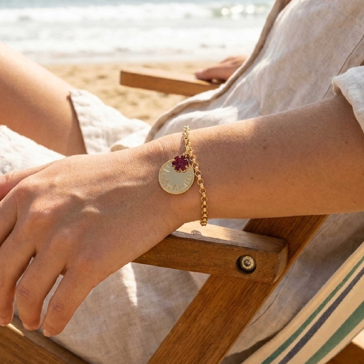 A gold chain bracelet with a round gold medical ID medallion and a small red star of life charm, worn on a woman's wrist in a beach chair at the beach.
