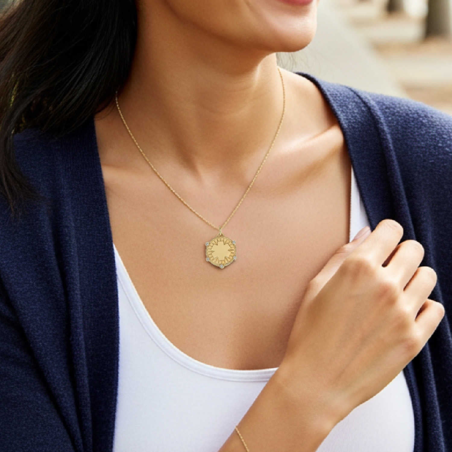 A close-up of a woman wearing a delicate gold necklace with a textured, circular medical ID medallion pendant. She is wearing a white tank top and a navy blue cardigan, with her hand resting near her chest.